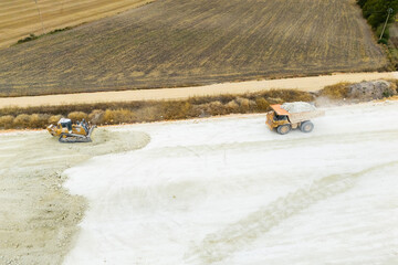 An aerial view of heavy machinery including a dump truck unloading soil, a bulldozer, and a grader working on a road construction embankment. © scharfsinn86