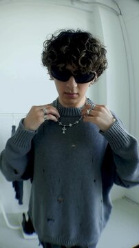 Young man in grey sweater adjusting cross necklace with sunglasses indoors