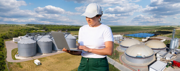 A female engineer using a laptop computer against a panoramic aerial view of an industrial complex with grain silos and biogas fermenter domes © scharfsinn86