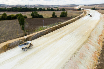 An aerial view of a highway under construction stretching across rural fields with a loaded dump truck driving on the dirt embankment. © scharfsinn86