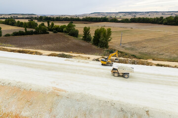 An aerial view of a highway construction site with a yellow excavator and a loaded dump truck operating on a dirt embankment. © scharfsinn86
