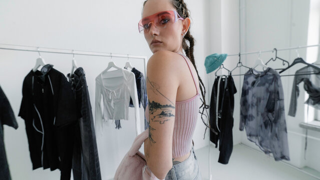 Fashion model with pink sunglasses posing among clothing racks in white studio