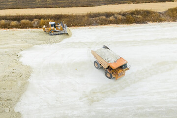 An aerial view of heavy machinery including a dump truck unloading soil, a bulldozer, and a grader working on a road construction embankment. © scharfsinn86