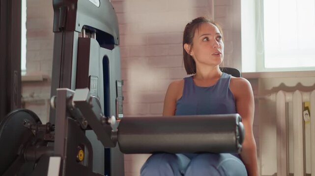 Woman using gym equipment for exercise. Woman is seated and interacting with weight machine in indoor fitness setting. Bright natural light fills room highlighting details of both subject