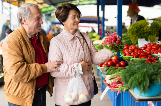Positive mature couple in casual wear picking fresh radish during shopping at crowded bazaar