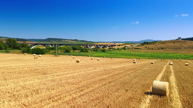 A bale rests in the foreground of a harvested Slovak field. Gold rows lead past scattered cylinders toward a distant viaduct below a broad blue sky.