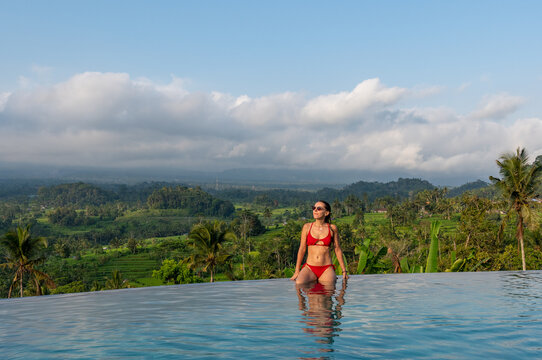 Bali, Indonesia - 05.10.2023: Woman wearing red bikini relaxes in an infinity pool with tropical valley landscape.