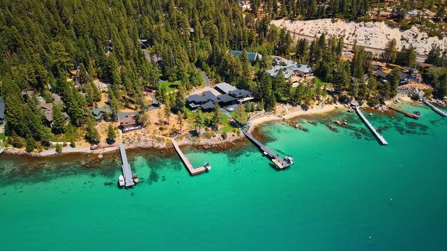 Cottages and docks line a forested shore at Lake Tahoe. Gray roofs sit among green conifers above turquoise water with narrow piers in an elevated view.