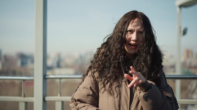 Woman with long curly hair wearing brown puffer jacket makes expressive hand gestures while speaking on outdoor balcony. scene features bright daylight and blurred cityscape in distance