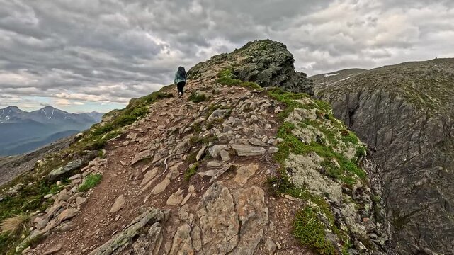 Person hikes rocky mountain ridge under cloudy sky. Trail winds steeply through exposed stone and low greenery. Lone figure pauses, taking in vast, misty valley view. Wind blows sparse trees Romsdalse