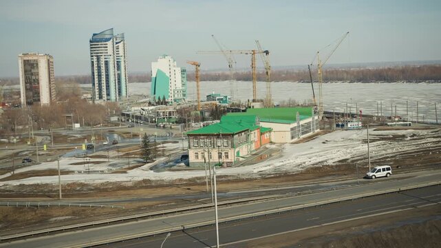 Panoramic view shows urban landscape with high rise buildings under construction near large body of water. Construction cranes dominate skyline while vehicles move along roads