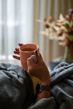 Person resting under a warm blanket and holding a cup of tea at home, representing a sick day, recovery, comfort and self-care.