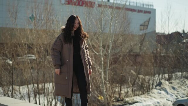 Woman strolls along outdoor path during winter. Woman wears lengthy padded jacket with fur trim over black clothing as bare branches of trees and patches of snow frame scene behind her