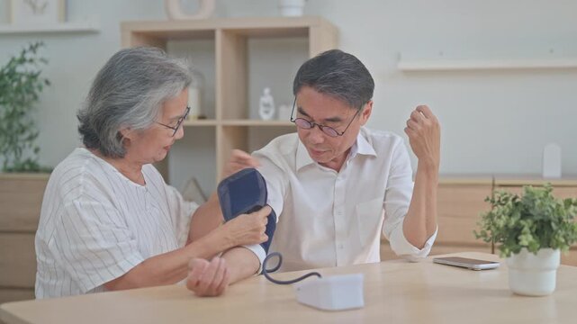 Elderly wife wraps a digital blood pressure cuff around her husband's upper arm at the table, checking his health during their routine at home.