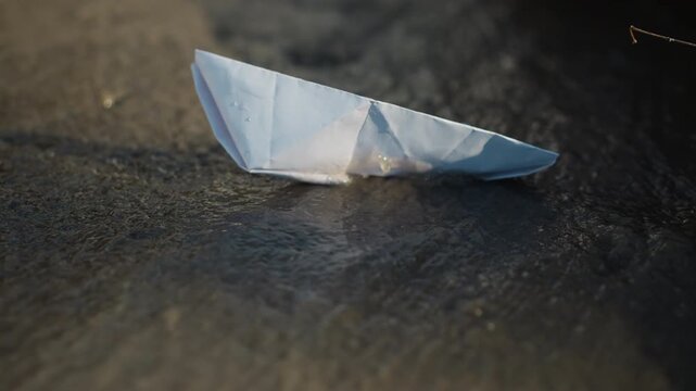 Small origami style folded piece of white paper lies stationary and slightly tilted upon damp ground. surrounding area features textured stone surface wet patches indicating recent