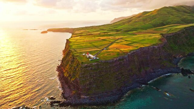Sunrise over Farol do Albarnaz lighthouse on northwest Tip of Flores Island, Azores, Portugal. Volcanic coast, patchwork green fields, Corvo island in distance, Atlantic ocean. Aerial view. Orbiting