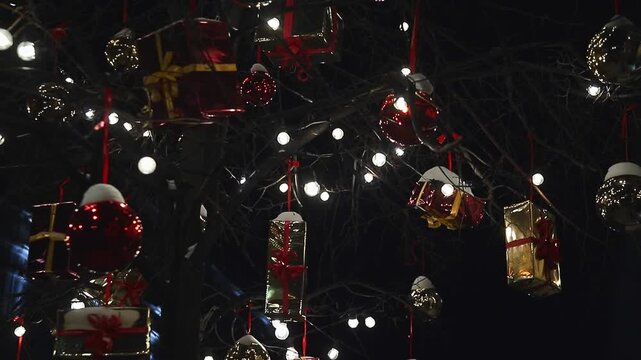 Tree decorated with glowing string lights, hanging gift boxes and red baubles at night. Festive Christmas holiday atmosphere