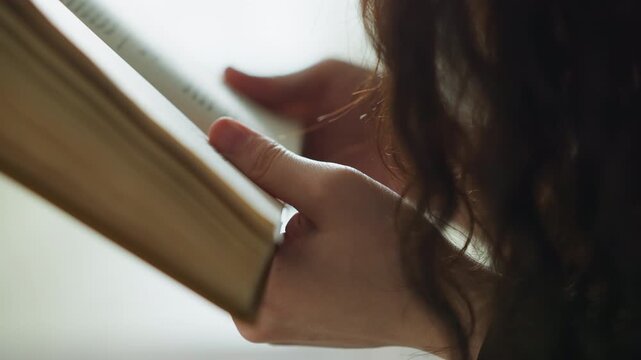 Close up shows woman hand gently flipping through thick hardcover book with visible text. Soft daylight illuminates page edges and fingers creating warm bokeh background