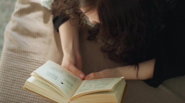 Woman with long hair lies down and holds opened book in her lap. Her fingers rest gently over pages as woman reads. scene is softly lit focusing on hands interacting with physical text