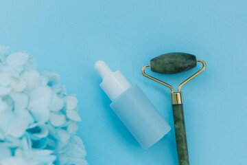 Cosmetic dropper bottle and green jade face roller placed with blue flowers on a plain light blue background © ir1ska