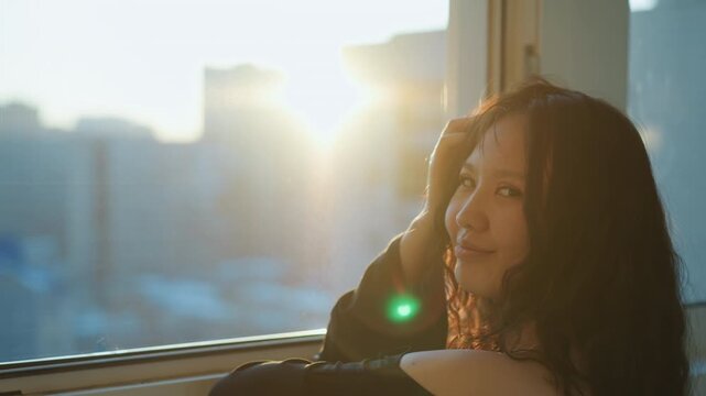 Woman gazes out window at sunset. Her profile is visible from behind as woman rests her hand on glass and adjusts loose strands of hair. Soft golden light filters through creating warm glow