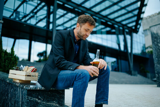 Jobless corporate employee holding alcohol bottle on steps deals with severe career failure.