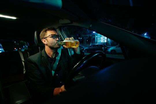 Drinking beer behind steering wheel inside vehicle at night causes dangerous traffic risks.