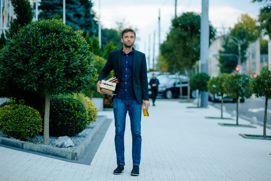 Unemployed corporate employee walks down urban sidewalk carrying office box after dismissal.