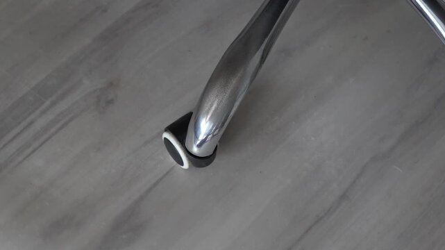 Chrome leg and black caster wheel of an office chair resting on grey wood effect laminate floor, close up showing modern furniture detail, concept of home office, ergonomics and floor protection