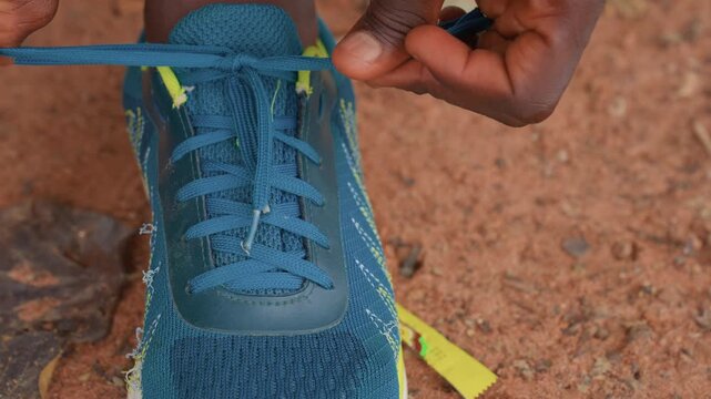 Hands carefully tie shoelaces on worn running shoe placed against earthy terrain. focus remains steady highlighting detailed hand movements over blue sneaker with frayed fabric near sole