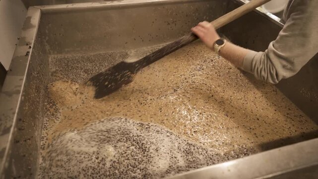 Worker stirs a large vat of foaming liquid with a long wooden paddle during an industrial mixing process in a stainless steel tank
