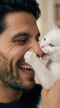 A close-up portrait of a laughing man while a small white kitten playfully bites his nose indoors, hilarious pet moment, cheerful lifestyle scene, adorable animal interaction.
