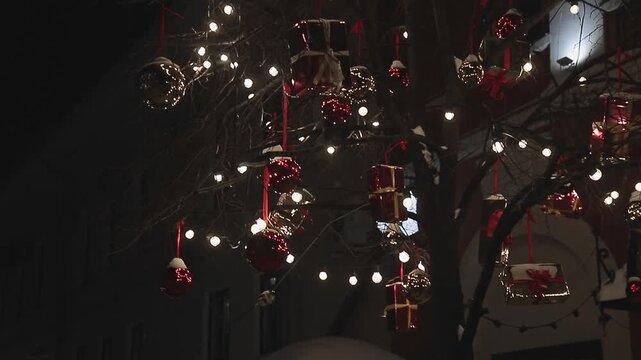 Tree branches adorned with glowing string lights, red baubles and gift box ornaments at night. Festive Christmas holiday decoration outdoors