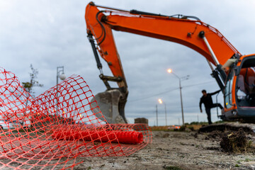 An orange hydraulic excavator lowers its bucket beside a worker at a muddy construction site, with red safety mesh in the foreground. © Marina