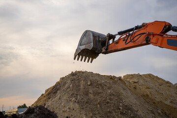 An orange hydraulic excavator arm with a toothed steel bucket hangs above a mound of excavated soil at a construction site under a cloudy sky. © Marina