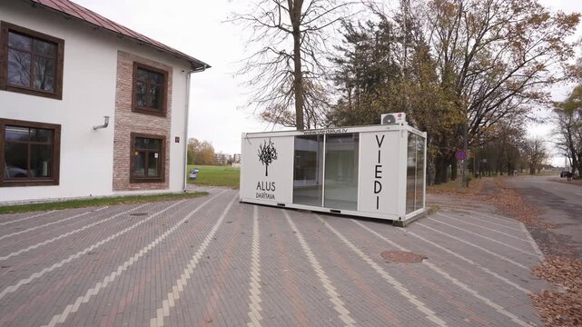 Exterior view of Alus Daritava Viedi beer brewery building and branded glass kiosk beside a quiet tree lined street in autumn