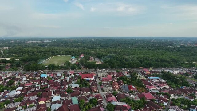 Aerial view of a quiet small town settlement with dense red-roofed houses, narrow streets, and lush green tropical nature surrounding the urban area under a bright daylight sky.