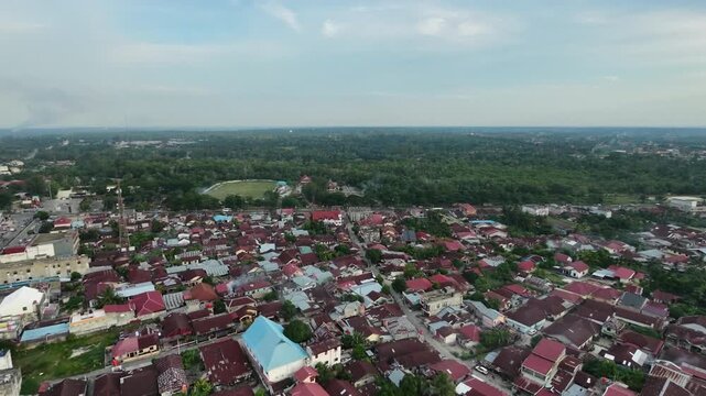 Aerial view of a quiet small town settlement with dense red-roofed houses, narrow streets, and lush green tropical nature surrounding the urban area under a bright daylight sky.