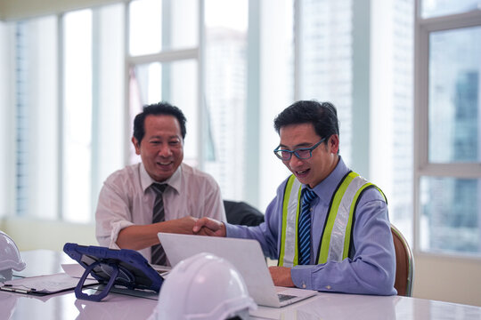Asian engineer construction engineer handshake project discussion office meeting safety vest laptop blueprint white helmet