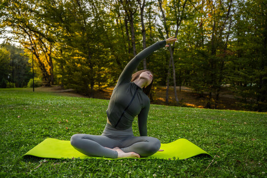 A woman performs a seated side stretch on a yoga mat in a grassy park, raising one arm overhead as autumn trees fill the background.