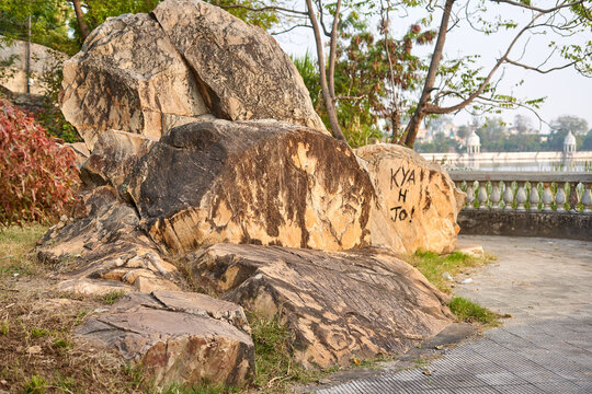 Large weathered boulders and rock formations occupy the foreground of this outdoor scene, situated beside a paved walkway. One prominent rock face features black spray-painted graffiti reading "KYA