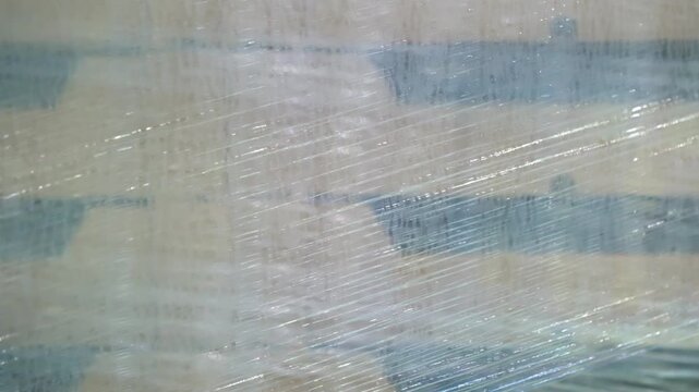 Cardboard boxes wrapped in stretch film. Stacked packages show blue stripe patterns. Tight plastic wrap covers each container. Blurred warehouse background visible