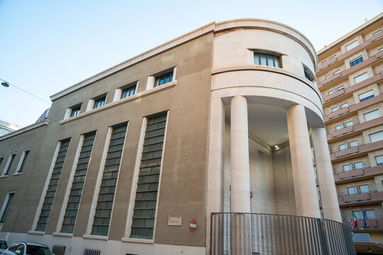 Bari, Italy - Jul 31, 2026: Rationalist era building with a curved corner colonnade and tall vertical windows on a Bari street, beige stone facade beside a modern apartment block under a clear blue s