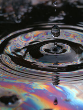 Clear water droplet falling onto iridescent oil film, creating circular ripple with vivid rainbow refraction and surface tension in dramatic macro lighting