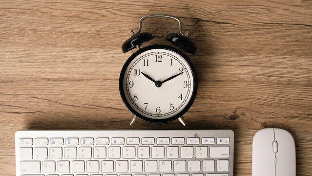 Black vintage alarm clock on wooden desk with computer keyboard and mouse representing time management deadline work hours remote work efficiency and productivity