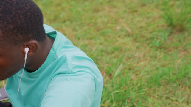 Young adult male rests outdoors in relaxed pose with arms behind head while listening through white earbuds. Man wears light blue t shirt against green grass suggesting casual downtime