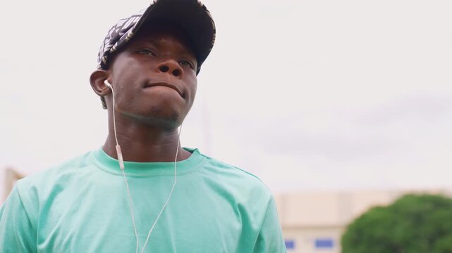 Shows young adult male listening to music or podcasts through white earbuds while standing outdoors under cloudy skies. Man wears patterned cap and light green t shirt with soft focus