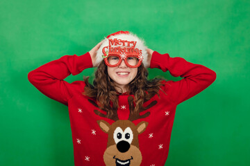 Young girl wearing festive red sweater with reindeer design and oversized glasses celebrating Christmas against a bright green background © Natalia