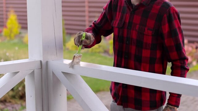 Homeowner painting a white wooden porch railing with a brush, performing exterior renovation and maintenance work outdoors.