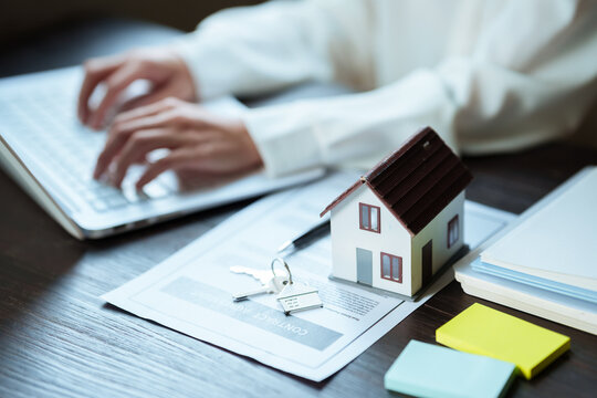 Real estate professional working on a laptop beside a house model, property contract, and keys, representing home buying and real estate transactions.